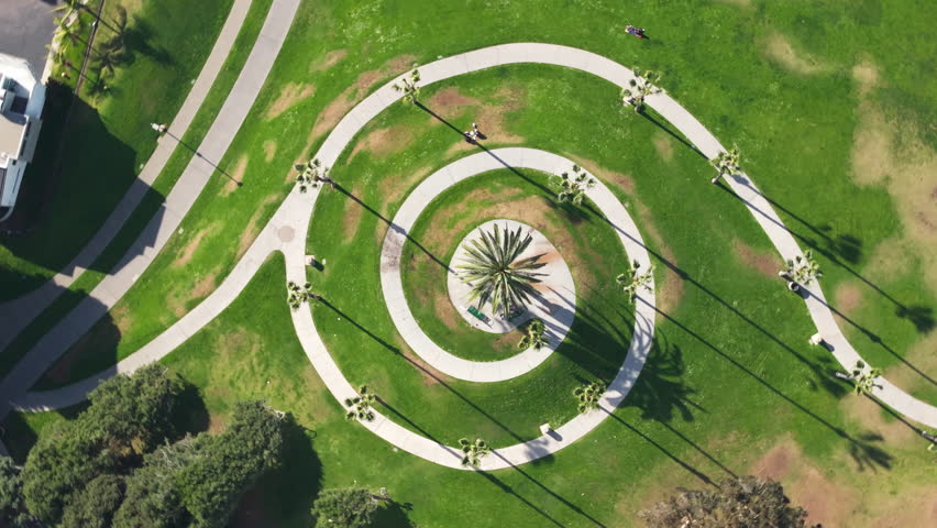 Aerial drone footage captures stunning spiral walking path design in Santa Monica California USA park with central palm tree casting long shadows on green grass lawn and curved concrete pathways.