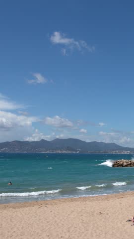 Wide static shot of a sandy beach, turquoise ocean, and mountains under blue skies.