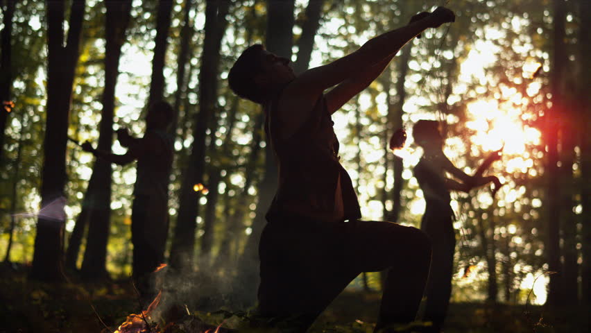 Silhouette performers spin flaming poi in a forest at sunset, artistic ultra slow motion fire performance.