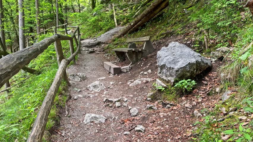 Picnic table and wooden railing along a rocky forest trail, surrounded by green woodland, boulders, roots, and peaceful hiking scenery.