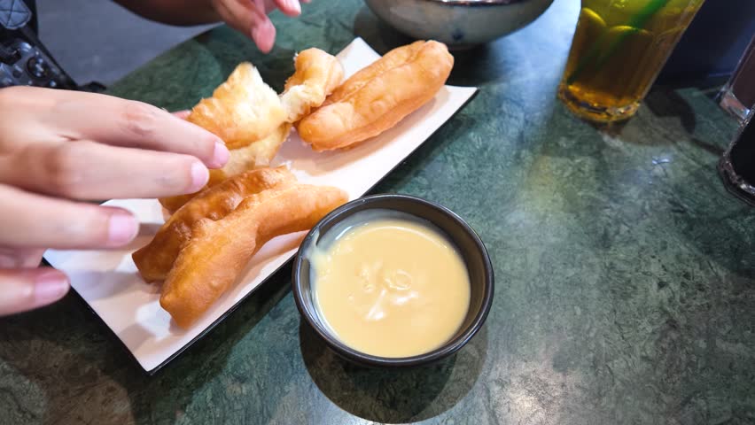 Close-up of hands dipping golden fried dough into creamy custard sauce on a marble table.
