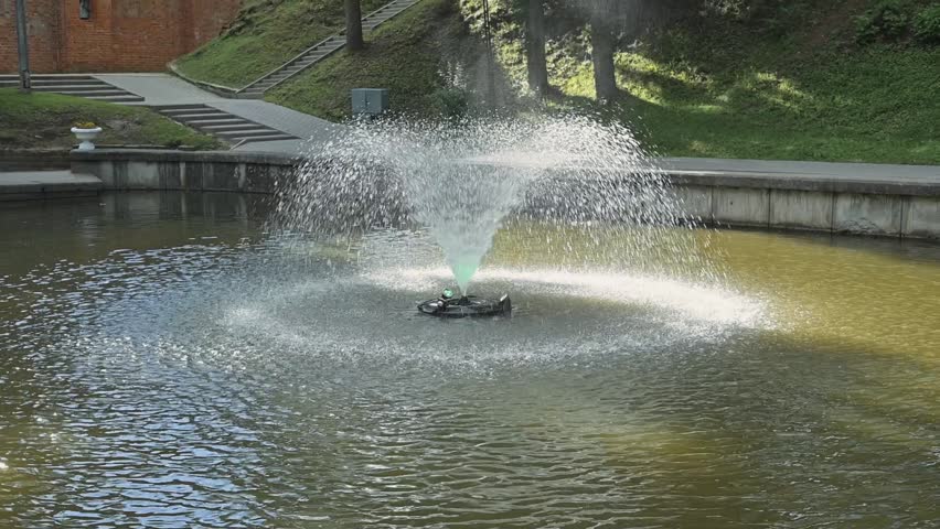 A water fountain sprays water into the air in a park surrounded by grass and trees. Sunlight reflects off the water creating a shining effect on the surface.