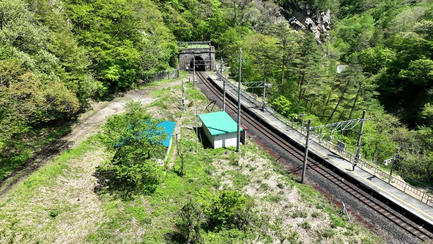 Aerial footage of an E721 Series local train arriving and departing Omoshiroyama-Kōgen Station on the JR East Senzan Line. Captured on a clear early summer day, the video follows the four-car commuter train through a beautiful mountain gorge surrounded by forests, waterfalls, and the historic Senzan Tunnel portal. One of the most scenic railway sections connecting Yamagata and Sendai.