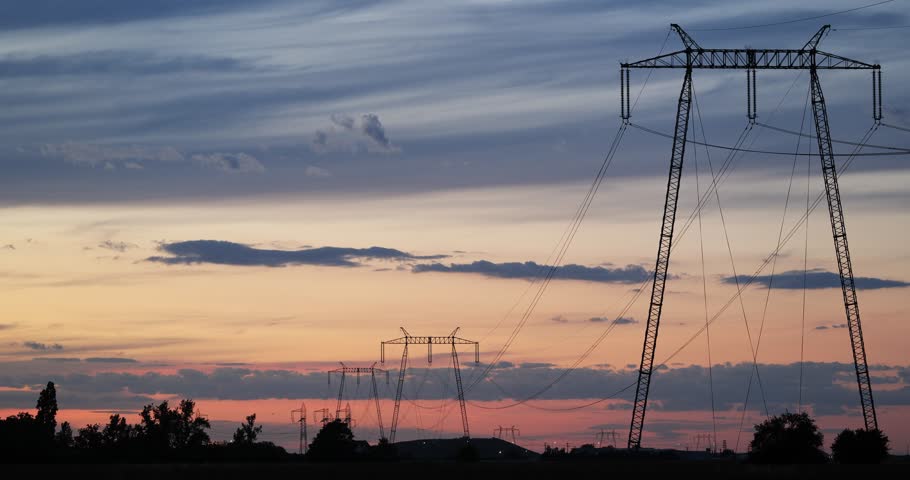 Wide shot of high voltage power lines and electricity transmission towers standing in a row as silhouettes against a beautiful orange and blue sunset sky during evening twilight.
