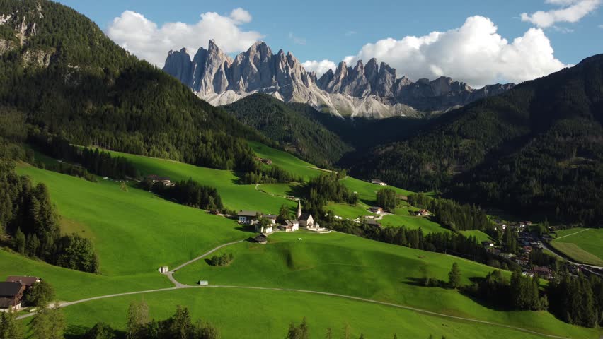 Aerial drone footage of the iconic Church of St. Magdalena surrounded by lush green alpine meadows in Val di Funes valley, with the dramatic Odle (Geisler) Dolomites peaks rising in the background, South Tyrol, Italy.