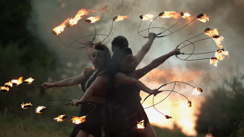 Three performers spin flaming props, forming a fiery halo in smoky outdoor ultra slow motion.