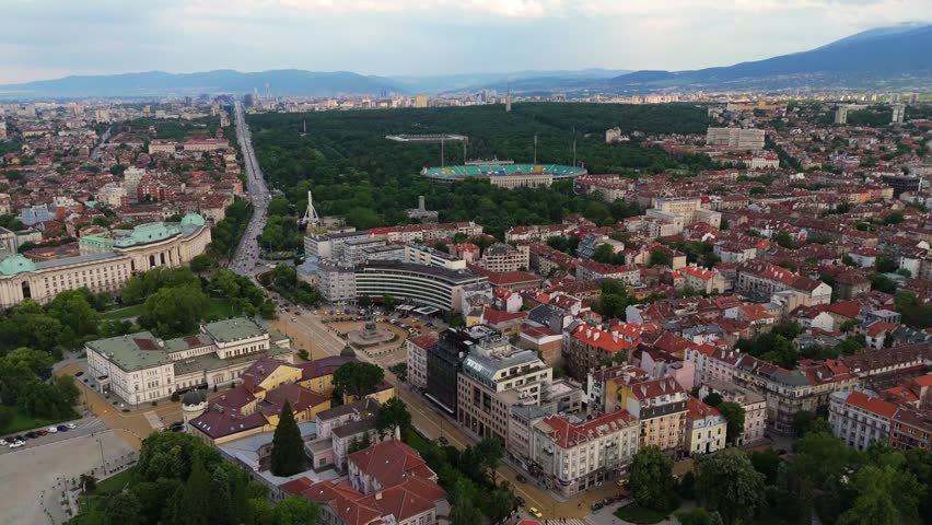 wide aerial view overlooking the historic city rooftops and green urban parks of Sofia, Bulgaria. Daytime drone cityscape tracking shot under a clear sky.
