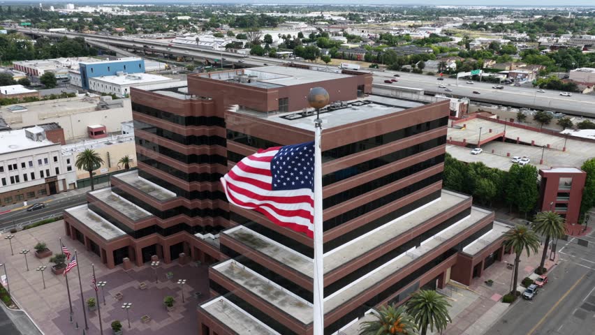 Patriotic American flag waves atop striped high-rise government building in downtown Stockton, California, cars traveling nearby freeway beside sunny palm-lined civic center plaza. Aerial drone orbit.