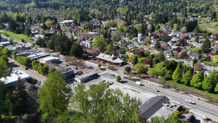 Historic railway depot anchors charming Snoqualmie, Washington town nestled among evergreen forest, tree-lined streets winding past houses beneath forested mountains. Aerial drone flyover shot