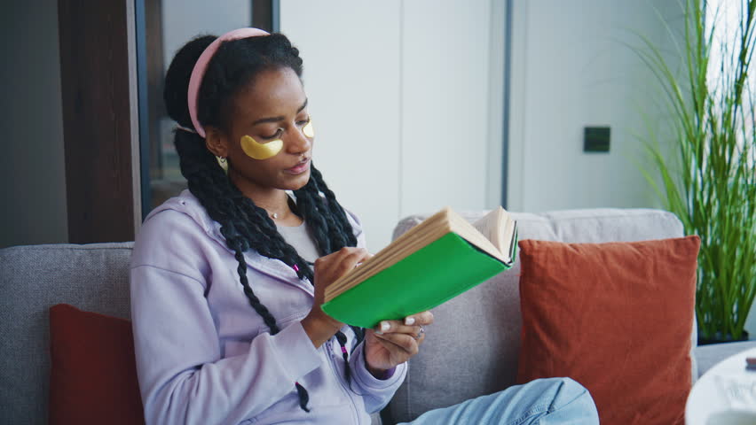 Person reads book on couch while wearing eye patches and relaxing at home during daytime