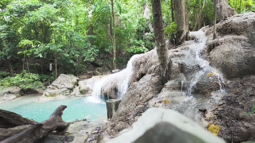 Clear stream water slips over textured rock tiers into an aqua pool in a lush Kanchanaburi woodland, with a gentle rack focus shift.