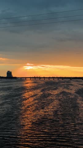 Scenic sunset over a wide river with the sun setting directly over a long bridge. Bright golden reflection on the rippling water surface under moody clouds. Calm evening landscape.