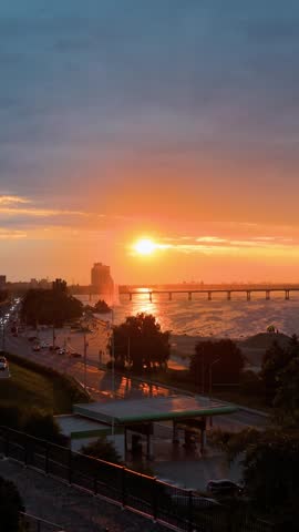 Spectacular high angle view of a bright golden sunset over a city river. Radiant sun illuminates the embankment road with traffic, a water fountain, and a long bridge.