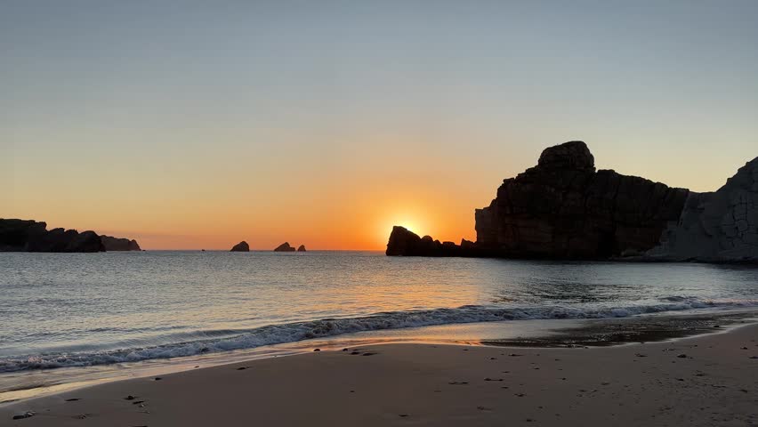 Golden sunset as the sun slowly sets behind a rocky islet, illuminating a calm rocky cove with gentle waves and warm evening light.