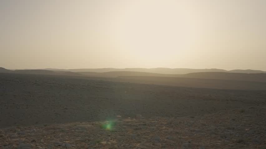 Sunset glows over the vast desert landscape. Low hills fade into the hazy horizon. Green lens flare appears in the foreground. Empty sandy terrain stretches to the evening sun. Ramon Crater in Israel