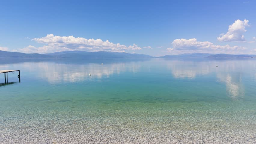 Drone flies low over the calm waters of Lake Ohrid with mountain reflections.
