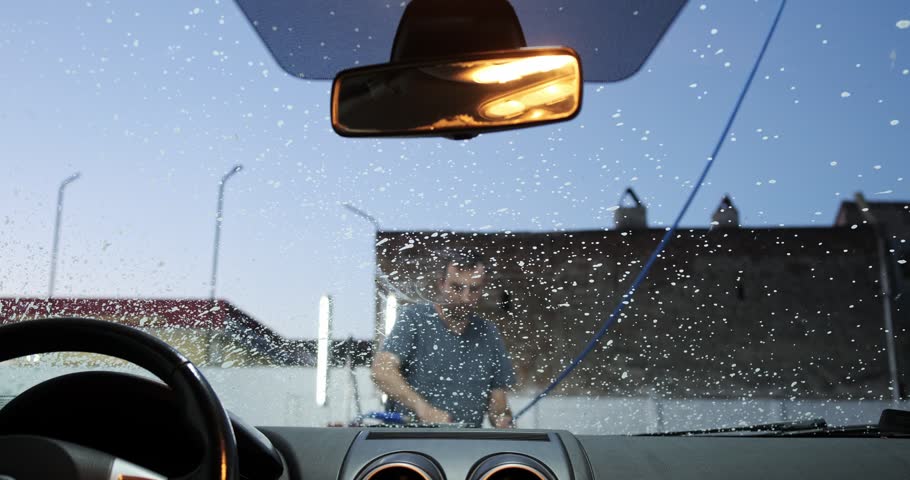 Point of view shot from inside a vehicle dashboard looking through the windshield as a man uses a pressure washer lance to spray thick white cleaning foam soap during a self-service car wash.