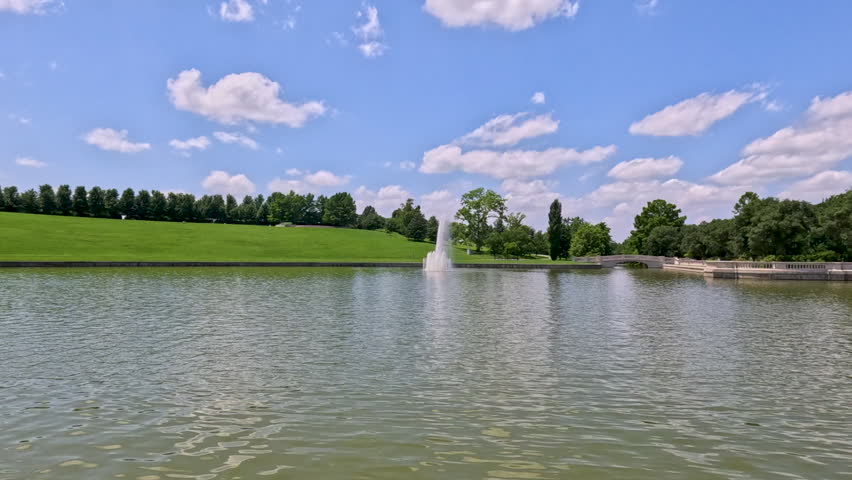 Scenic Park Fountain and Historic Stone Footbridge. Slow-motion views capturing a classic grand park water fountain feature with a detailed stone walking bridge in the background on a sunny day in Forest Park, St. Louis, Missouri, USA.