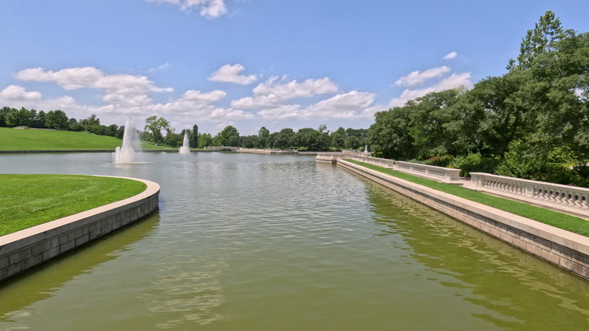 Elegant Water Fountains and Grand Basin Landscape. A beautiful wide shot showcasing water fountains spraying and blowing in the wind inside a large public park basin, bordered by an ornate stone banister and a grassy hillside at Forest Park in St. Louis, Missouri, USA.