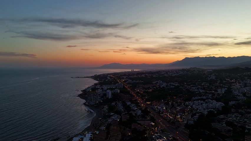 Hyperlapse aerial drone footage of dusk over the coast near Mijas, Costa del Sol, Spain, with a coastal highway, car lights, calm sea, and mountain silhouettes