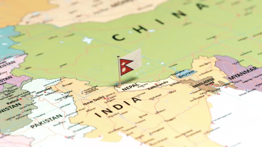 A high-angle macro photograph focusing on a printed political map of South Asia, specifically highlighting the landlocked Himalayan nation of Nepal. A miniature national flag pin of Nepal, displaying its unique double-pennon shape with a red background, blue border, white crescent moon, and sun symbols, is placed securely into the map territory near the capital city of Kathmandu. Surrounding regions and neighboring countries—including India to the south, China (Tibet) to the north, Bhutan, Bangladesh, and Pakistan—are visible on the detailed map layout with a soft background blur and shallow depth of field. This professional composition is ideal for Nepal tourism promotions, Himalayan travel vlogs, geographical education, regional news, and international business concepts.