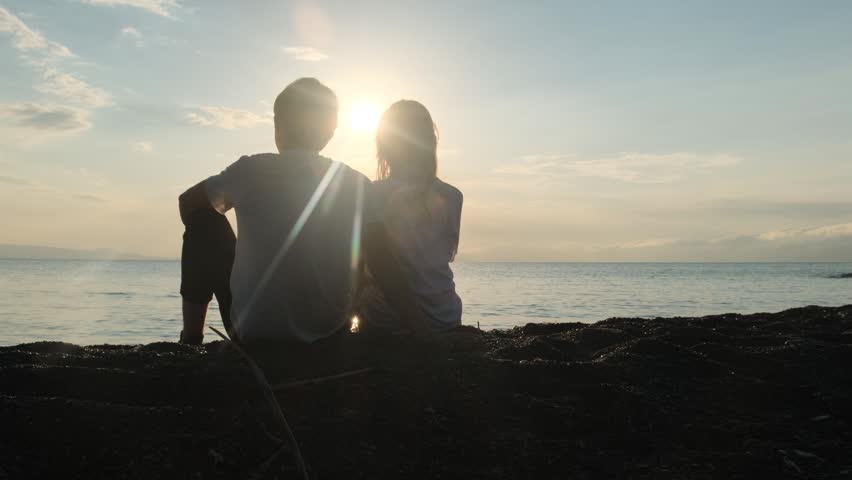 Young couple, a teenage boy and girl, sit shoulder to shoulder on the seashore watching the sunset. Girl rests her head on the boys shoulder. First love, teenage romance, romantic relationship.