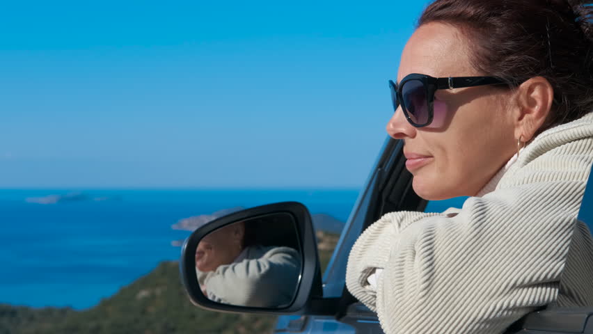 Woman admiring ocean view during car road trip. Serene woman wearing sunglasses enjoys a scenic coastal drive, looking out the car window at the beautiful blue sea and green hills