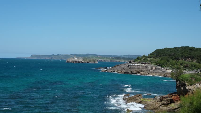 Panoramic seascape with a distant horizon. Turbulent, dramatic crashing waves into a rocky shore. Lighthouse on the little island. Santander, Cantabria, Spain. Tourist routes concept.