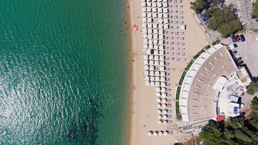 Straight-down drone view of a beautiful beach near Kavala, Greece, showing turquoise water, sandy shoreline, neat rows of parasols and sun loungers, and a curved beachfront building.