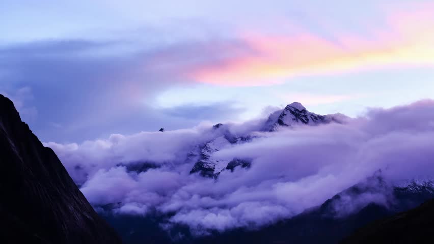 Breathtaking view of the snow-covered peaks of Makalu Barun National Park in eastern Nepal, captured during the tranquil blue hour. Dramatic clouds drift around the rugged Himalayan mountains, creating a spectacular alpine landscape with a peaceful and cinematic atmosphere. This stunning natural scenery is ideal for travel, adventure, mountaineering, nature documentaries, environmental conservation, and Himalayan landscape productions.