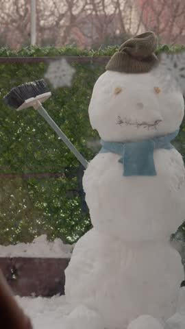 Vertical Screen: A boy stands by the window and watches a snowman in the yard. The snowman has a hat and scarf and looks fun. Snow is all around, creating a winter scene.