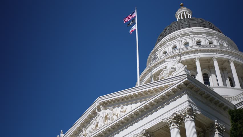 California State Capitol is shown in Sacramento with bright sun and blue sky. The flag is waving above the building.