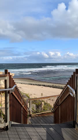 Stairs lead down to a sandy beach and ocean with waves. Clouds are in the sky above the water.
