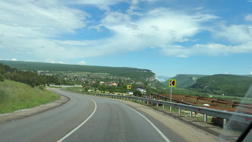 Scenic asphalt road through rural landscape with hills. Front facing dashboard camera shot driving towards a small village on a sunny day.