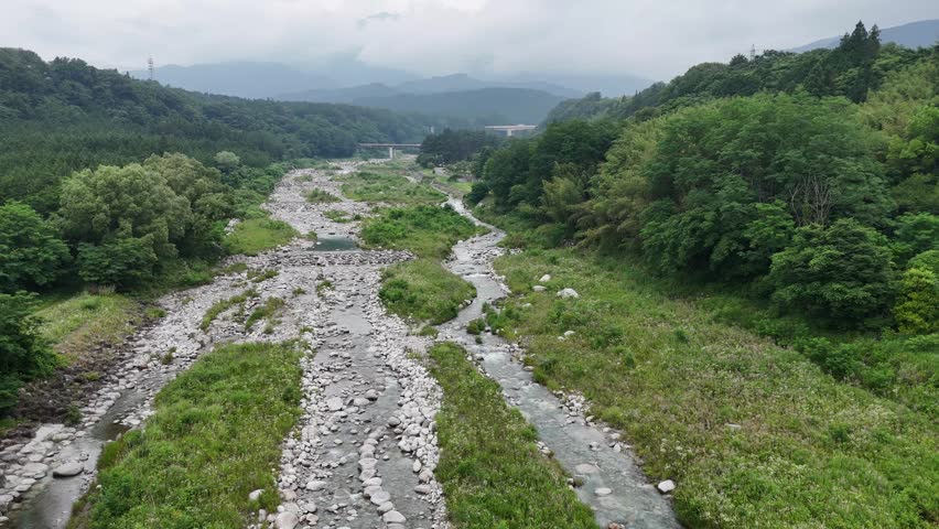 Riverbed flows through lush green valley. Rocks and grass surround the waterway. Clouds hang low over distant hills. Nature
