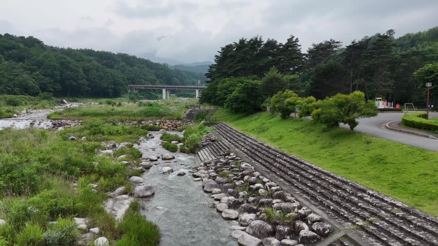 River flows gently through green, rocky banks. Clouds hang low above forested hills and a distant bridge. Grassy slopes border the stream, edged with stone retaining walls