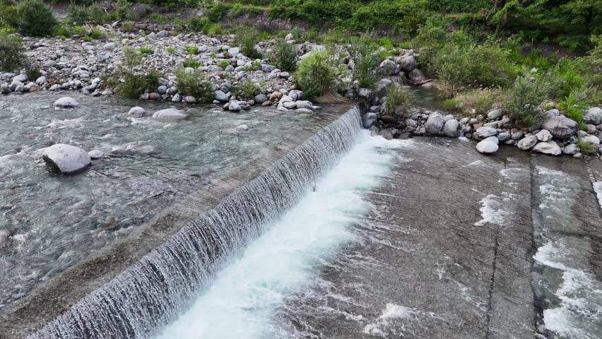 Clear water flows over a simple concrete dam. Rocks line the riverbed, surrounded by greenery. Foam swirls as water cascades down the stepped drop. Nature