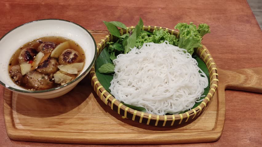 Bun Cha is a classic Vietnamese dish featuring grilled pork served in a savory-sweet dipping broth with pickled vegetables. In this image, a bowl of grilled pork slices and meatballs in broth is accompanied by a plate of fresh rice vermicelli noodles, lettuce, and aromatic herbs.