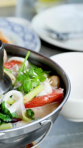 Close-up of ladle serving spicy seafood soup from a metal hot pot into a bowl.