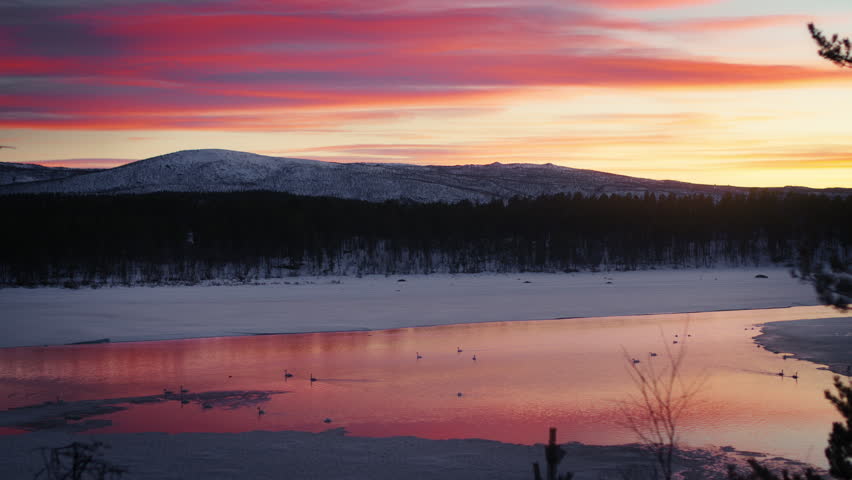 Colorful sunset clouds reflect on a partly frozen river below dark forest and snowy hills. Ideal for nature, winter landscape, travel and background content.