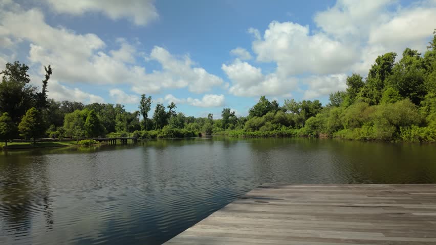 Wide view of a calm freshwater lake reflecting trees and clouds on a sunny day. Beautiful natural landscape with lush shoreline, walking paths, and tranquil outdoor scenery.