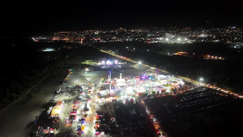 Drone performs a right orbit with a slight pull in over the Mexican fair in San Jose del Cabo, revealing the fairground, parking lot traffic below, and glowing town lights stretching into the distance