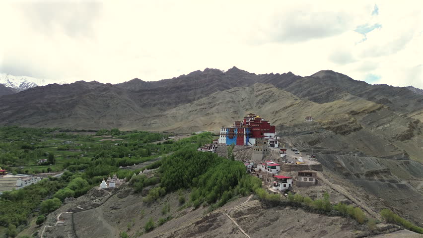 High-angle drone pan shot revealing a wide panorama of Stakna Buddhist Monastery on a hill, surrounded by white chortens, a green valley, and massive barren mountain ranges in Ladakh, India.