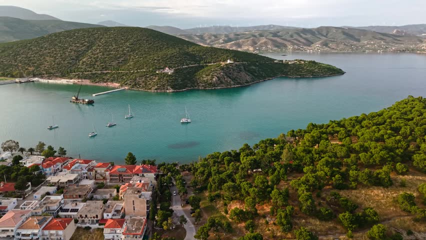 Wide drone shot of Ermioni, Greece at golden hour. Lush green pine forest bordering a calm turquoise sea bay with anchored yachts and mountains