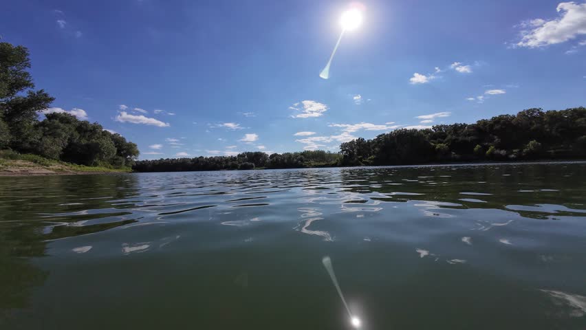 POV shot of a boat moving fast on a wide river under a blue sky with sun reflections