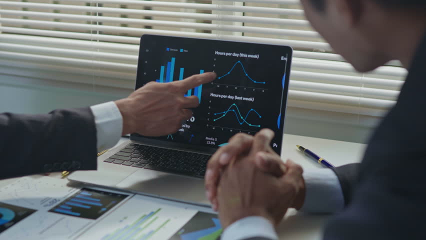 Business professionals discussing and explaining data charts on a laptop screen during an office meeting, with reports and documents on the desk, representing teamwork, analysis and strategic planning.