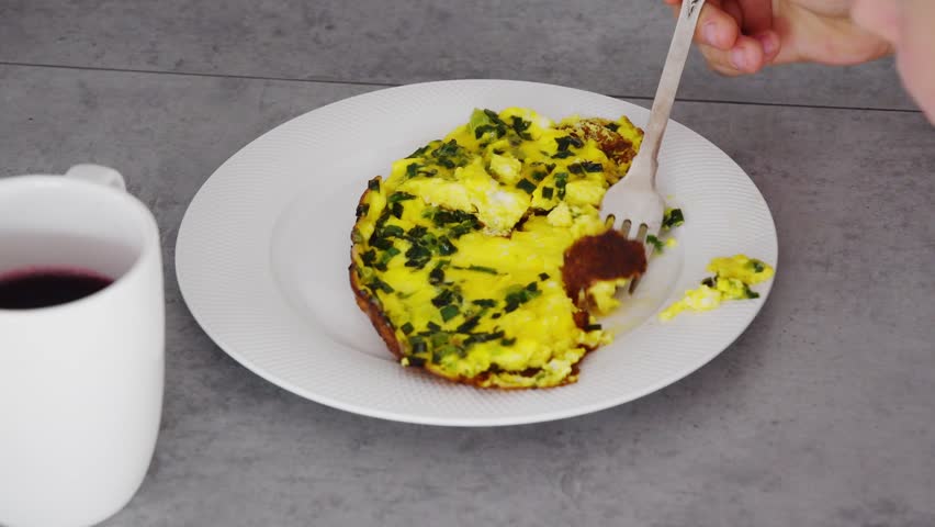 High angle view of a person using a fork to eat a delicious egg frittata with chives from a white plate, enjoying a healthy homemade breakfast served with a cup of red grape juice