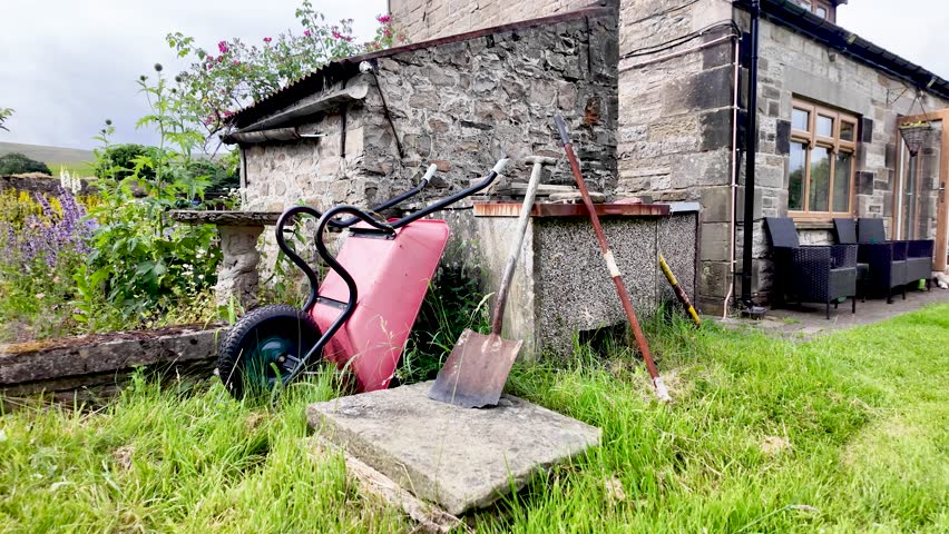 Rusty wheelbarrow and shovels rest in long grass beside an old stone house in a quiet rural yard.