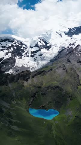 An aerial view captures a stunning turquoise glacial lake surrounded by majestic snow-capped mountains and rocky green slopes.