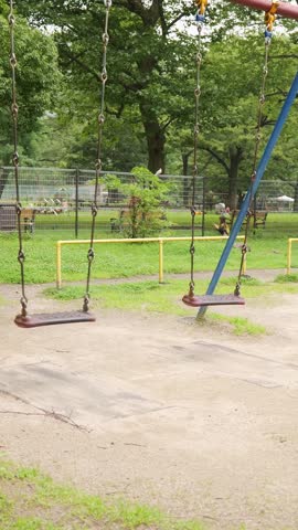 Empty chain swings gently swaying in a quiet urban park surrounded by green trees and grassy ground on a calm daytime.